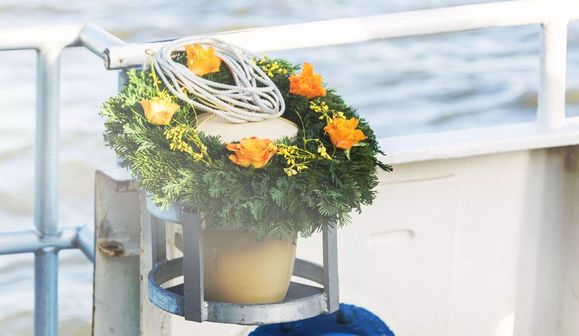 Urne mit Blumenschmuck auf einem Schiff: Eine helle Seeurne steht in einer Halterung am Geländer eines Schiffs. Sie ist mit einem Kranz aus Tannenzweigen und orangefarbenen Blüten sowie einem weißen Seil geschmückt. Im Hintergrund ist das Wasser zu sehen.